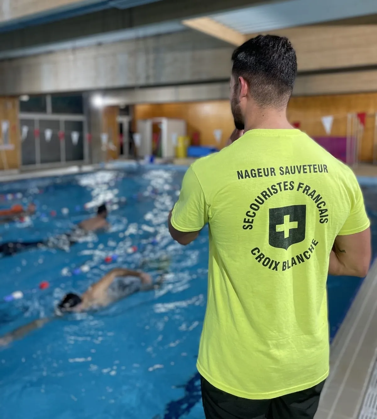 Un nageur sauveteur de dos, en tee-shirt jaune Secouristes Français Croix Blanche, surveille un bassin.