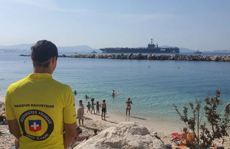 Un sauveteur en tee-shirt jaune, de dos, surveille une plage depuis les rochers.